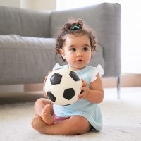 Adorable black haired baby girl in pale blue clothes sitting on floor at home, looking away, playing soccer ball. Kid at home and childhood concept