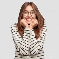 Indoor shot of happy beautiful student keeps hands under chin, glad to recieve praise, wears round spectacles, spends free time with close friends, stands against white background. Happiness concept